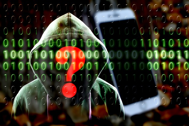 Image of hooded person masked behind binary code with red question mark obscuring his/her face, they are standing next to a smart phone.